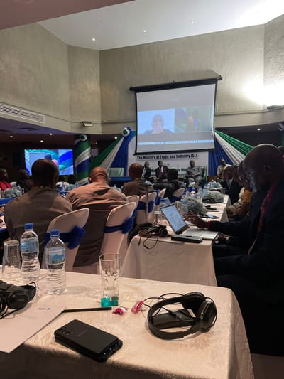 This image captures an indoor meeting or conference taking place in Freetown, Sierra Leone, likely during the daytime under artificial lighting. The scene shows multiple individuals, predominantly men, seated at long tables covered with white tablecloths. The chairs are adorned with white covers and distinct blue bows, matching blue decorative elements on the tables.

In the background, a large projector screen displays a video call, showing two individuals, indicating remote participation or a hybrid event setup. Below the screen, a banner is visible with the prominent text: "The Ministry of Trade and Industry (SL)". Partially visible text beneath this includes "Directorate of" and "Ministers of", strongly suggesting a government or ministerial meeting related to trade and industry in Sierra Leone. The draped fabric in the background features blue, white, and green colors, consistent with Sierra Leone's national flag.

Attendees are seated facing the front of the room, with some using laptops or tablets placed on the tables. Each place setting includes bottled water, clear glasses, and in some instances, headphones, implying simultaneous translation or audio equipment. In the foreground, on a table to the right, a pair of black headphones and a black smartphone are visible, along with another water bottle and a clear glass. A document with "Radisson Blu" is also faintly seen on one of the tables, possibly indicating the venue or a sponsor. The atmosphere appears professional and focused, with participants engaged in the proceedings of the event.