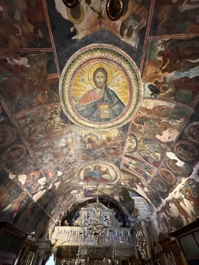 The image shows the interior of a church in Kassopaia Municipal Unit, Greece, with a frescoed ceiling depicting religious scenes. A central fresco shows Jesus Christ, surrounded by a halo and radiating light. Other frescoes around the ceiling depict various figures, possibly saints or biblical characters, engaged in different actions. The artwork appears aged and somewhat faded, with some areas showing signs of wear and tear. Below the ceiling, an ornate chandelier hangs, and parts of the church's altar or iconostasis are visible, adorned with intricate carvings and religious icons. The overall impression is one of a historic and sacred space.