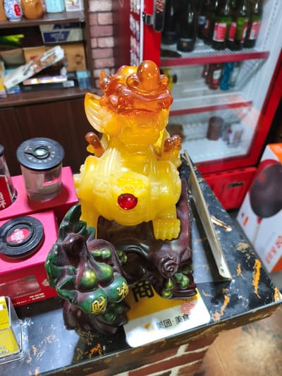 A close-up, eye-level shot captures a decorative statue of a mythical creature, likely a Pixiu or Qilin, made of translucent yellow material, possibly resin or amber, with orange translucent accents on its head and paws. A red gem is set into the creature's chest. The statue is mounted on a dark, carved base resembling a gnarled tree branch. In front of the statue, to the left, is a dark green carving of gourds with gold Chinese characters inscribed on them.

The statue and carvings rest on a black and gold speckled surface, possibly a tabletop or counter. To the left of the statue are several small, cylindrical containers with black lids. One is clear and contains a grey substance, and another is red with a black lid and Chinese writing. Behind these containers, a portion of a red refrigerator is visible, with shelves containing bottles of drinks. In the background, shelves hold various items, including boxes and decorative vases. The overall scene appears to be an indoor setting, possibly a shop or a home. The image does not provide any specific location within Jixi, China, other than suggesting an indoor commercial or residential environment. There are no people visible in the image, and no discernible actions or interactions are taking place. The lighting is moderate, illuminating the statue and its surroundings.