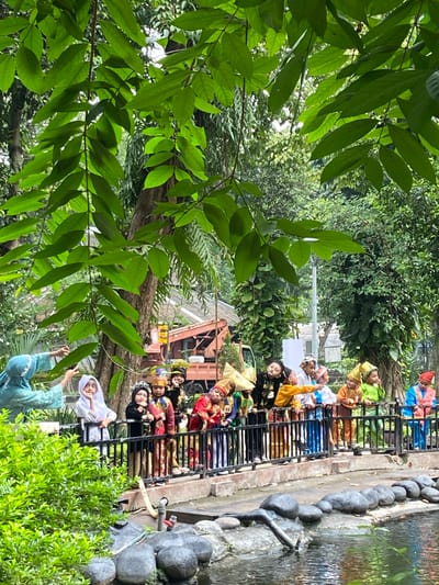 The original image is rotated 90 degrees counter-clockwise. When viewed upright, it depicts an outdoor cultural event in Surabaya, Indonesia, characterized by lush greenery and a water feature.
The central subjects are a group of young children lined up behind a black metal fence, which borders a shallow water body with a rocky edge and a small pipe extending into it. These children are dressed in vibrant, traditional Indonesian attire, featuring rich colors such as gold, red, blue, and green, often complemented by ornate hats or headbands. Their expressions and postures vary; some are actively observing something to their right, while others appear more relaxed, with one child in a black and gold outfit seemingly asleep or resting on the fence. The number "51" is visible on one child's garment.
In the foreground, large, verdant leaves from an overhead tree branch frame the scene. The background is filled with dense green foliage, through which a faint structure, possibly part of a building or an old vehicle, and a white utility pole can be discerned. Two adults are also visible: one in the lower foreground wearing a light blue traditional dress and hijab, extending an arm, and another in a white hijab standing further along the fence.
The scene unfolds during bright daytime under clear weather conditions, suggesting a school outing, cultural performance, or local festival. The overall atmosphere conveys a sense of vibrant cultural engagement within a serene natural setting.