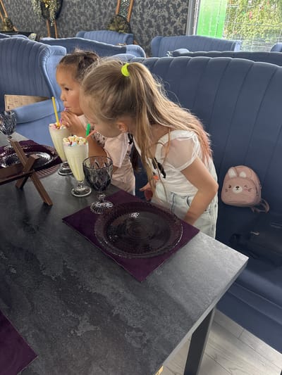 Two young girls are sitting at a table in an indoor establishment, likely a cafe or restaurant, in Anapa, Russia. The girl on the left, with dark hair in a bun, is drinking a milkshake with a yellow straw. The girl on the right, with blonde hair in a ponytail, is leaning over and also appears to be drinking a milkshake, possibly with a green straw. Both girls are wearing light-colored tops. On the table are two glasses filled with milkshakes topped with marshmallows, a dark glass, and two dark plates with purple placemats. A small pink backpack with a bunny design is visible on the blue upholstered bench seating behind the girl on the right. The background shows more blue seating and decorative elements. The floor appears to be light-colored wood laminate.