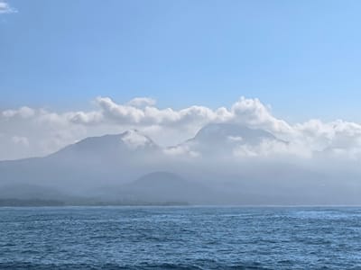 The image depicts a vast expanse of the ocean in the foreground, with ripples and waves suggesting gentle movement under a bright sky. In the background, a chain of mountains rises, partially obscured by soft, white clouds that drift around their peaks. The sky is a clear, pale blue, with a few scattered clouds higher up. The overall scene is serene and natural, evoking a sense of distance and tranquility. The location is unknown in Indonesia.