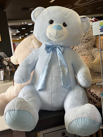 This media file shows several large, plush teddy bears displayed in a retail environment. The main subject is a light blue teddy bear positioned centrally, with a light blue satin ribbon tied around its neck. It has dark brown button eyes and a light blue embroidered nose and mouth. To the left and right of the blue bear are other teddy bears, one light brown and fuzzy, and another cream-colored. In the background, a heart-shaped pillow with the text "I LOVE YOU" is visible, along with more plush toys and retail displays. The setting appears to be a store aisle, with shelving and lighting visible. The overall impression is one of a merchandise display for stuffed animals. There is no indication of specific time of day, weather, or emotions. The text "I LOVE YOU" is clearly visible on the heart-shaped pillow. The location context provided is Studena, Bulgaria.