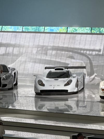 The image depicts an indoor exhibition space, likely a museum or showroom, featuring several sleek, high-performance sports cars. The cars are positioned on a glossy, reflective floor, creating mirrored images that extend the visual depth of the scene. A white, aerodynamic race car with a prominent rear spoiler is centrally located. To its left, another silver sports car with distinctive round headlights is visible. A portion of a third car, possibly white or light-colored, is seen on the far right. The background consists of a textured, illuminated wall with a pattern of horizontal lines and shadows, suggestive of a modern architectural design. Above this wall, a strip of windows reveals a blurred view of greenery and trees, indicating an outdoor setting beyond the enclosed exhibition space. The lighting is bright and diffused, highlighting the polished surfaces of the vehicles. The overall impression is one of a clean, well-maintained display focused on automotive excellence. This exhibition space is located in Stuttgart, Germany, as evidenced by the presence of the Porsche Museum, which is renowned for its collection of iconic sports cars.