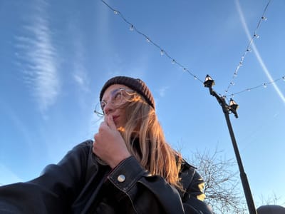 A low-angle outdoor shot captures a young person from below, appearing upside down against a clear blue sky. The person has long blonde hair, which is illuminated by sunlight, and wears a dark beanie, round glasses, and a black leather jacket. Their face is partially obscured by their hand.
In the upper left background, the intricate, bare branches of a tree are visible, suggesting a colder season like late autumn or winter. A tall, dark pole or lamppost runs vertically on the left side of the frame, from which several strands of decorative string lights with small clear bulbs are strung across the sky. Two antique-style street lamps are also suspended, contributing to an urban or park setting.
The sky is bright blue with faint, wispy clouds, indicating a clear, sunny day. The unique perspective suggests the person is looking down towards the camera while posing for a photograph. No text is visible in the image.