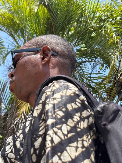 A male individual with dark skin and a close-cropped haircut is depicted in profile, facing left, outdoors in Igboefon, Nigeria. He wears black-framed sunglasses pushed up onto his forehead, with the arms resting above his ears. His attire includes a dark-colored garment patterned with irregular, light-colored shapes. A dark bag strap is visible over his right shoulder. The background features bright green palm fronds against a clear, light blue sky, indicating daylight conditions.