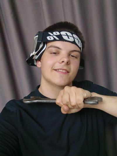 A young man with short brown hair is centered in the frame, holding a wrench to his neck with his right hand. He is wearing a black T-shirt and a black headband with white lettering, appearing to be goggles pushed up onto his forehead. His expression is a slight smile. The background is a plain, gray curtain. The scene appears to be indoors.
The location is Andreyevo, Russia.