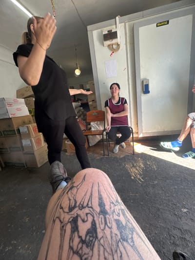 The image depicts a group of people in what appears to be a warehouse or storage area. In the foreground, a person's tattooed leg is visible, with a gray sneaker on their foot. To the left, a woman in a black top and pants is gesturing with her hands. Further back, another woman wearing a maroon vest over a black shirt and pants sits on a metal chair. Behind her, a large white door with the word "ВЫХОД" (Exit) in yellow lettering is visible. Stacks of cardboard boxes and bags are present throughout the space. The floor is concrete, with some areas of shadow and sunlight. The overall setting suggests a functional, possibly industrial, environment within Makiivka, Ukraine.