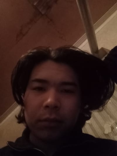 A low-angle selfie captures a young adult male, appearing to be of Central Asian descent, looking directly at the camera with a neutral expression. He has medium-length dark hair styled loosely, and is wearing a dark, possibly black, zip-up top with a visible silver zipper. The individual occupies the lower half of the frame. The background consists of a ceiling or upper wall area, illuminated by dim, uneven lighting. The upper left portion shows a dark brown, textured surface, possibly wooden paneling or patterned wallpaper. The upper right reveals a lighter beige or cream-colored surface with faint vertical stripes, along with a diagonally positioned light-colored structural beam or support. No specific identifying markers for Takhtakuprik, Uzbekistan are visible in the image itself, but the scene is situated within that city as per instruction.