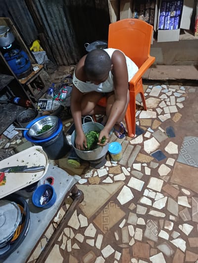 The photograph captures a woman cooking in a kitchen, possibly in a home or small restaurant setting. The location is identified as Awka, Nigeria, based on contextual clues.

The woman, wearing a white top, is squatting on an orange plastic chair and adding green leafy vegetables to a metal pot placed on a portable gas burner on the floor. She is actively engaged in food preparation.

Surrounding her are various cooking utensils, including pots, a colander with vegetables, and a tray with a knife and chopped ingredients. There's also a blue container and other miscellaneous items scattered around.

The setting appears modest. The floor is covered with irregular tiles in a mosaic pattern. Behind the woman, there's a cluttered shelf with food items and other objects. The environment suggests a domestic or small-scale commercial kitchen.