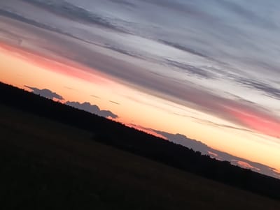 The image depicts a landscape view from the city of Chumna, Russia, during sunset. The sky is filled with a gradient of colors, transitioning from a soft pink and orange hue near the horizon to shades of gray and purple higher up, with wispy clouds stretched across. A dark silhouette of a forest lines the horizon, separating the sky from the foreground. Below the forest, a field is visible, predominantly dark and appearing to be covered in grass or low vegetation, hinting at an agricultural or natural area. The scene is captured at an angle, suggesting the photo was taken from a slight incline. There are no people or animals visible in the frame. The overall atmosphere is serene and tranquil, characteristic of a rural sunset.