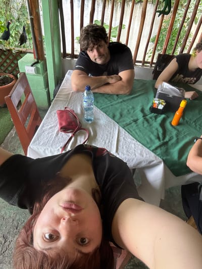 A selfie of a woman with brown hair and eyes is taken at a table. A man sits beside her, with crossed arms and dark curly hair. A child is also at the table. The table is covered in white and green cloths. On the table are a water bottle, a red purse, condiments, and a black box. The outdoor setting has green vegetation and wooden slats. Two black birds are visible outside.