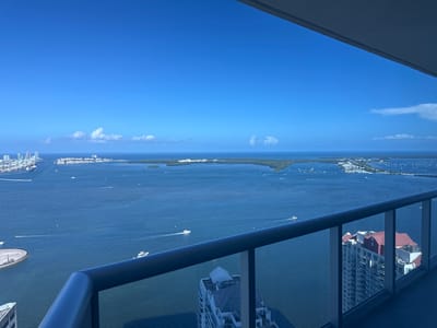 An aerial perspective captures a broad expanse of the Biscayne Bay, illuminated by a clear, bright blue sky with sparse, wispy clouds. The water is a deep, rich blue, dotted with numerous boats, ranging from small speedboats leaving white wakes to larger yachts. A distant city skyline is visible on the left, characterized by tall buildings and a port with cranes. The right side of the frame is dominated by a balcony railing, a dark blue metallic structure with glass panels. Beyond the railing, a distinctive white building with a red roof stands out. In the foreground, parts of other high-rise buildings are visible, suggesting the viewpoint is from a tall structure within the city. The overall scene depicts a vibrant coastal urban environment.