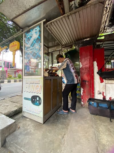 The image depicts a roadside key cutting and repair shop in Banjarbaru, Indonesia. A young man, wearing a baseball cap and a blue and white patterned shirt with the number "2" on the back, stands behind a counter and operates a key cutting machine. He is dressed casually in black pants and blue sandals. The shop is housed in a small, open-air kiosk with a corrugated metal roof and a glass front displaying images of keys and advertising text in Indonesian, including "Reparasi KUNCI" (Key Repair). A yellow sign with "WENXING KEY CUTTING MACHINES" is visible hanging from the kiosk's roof. To the right of the kiosk, there are large red and white painted columns, and a black and blue toolbox rests on the ground. In the background, a street with traffic and buildings can be seen, indicating an urban setting. The ground is paved with concrete, and a low concrete barrier is present in the foreground.
