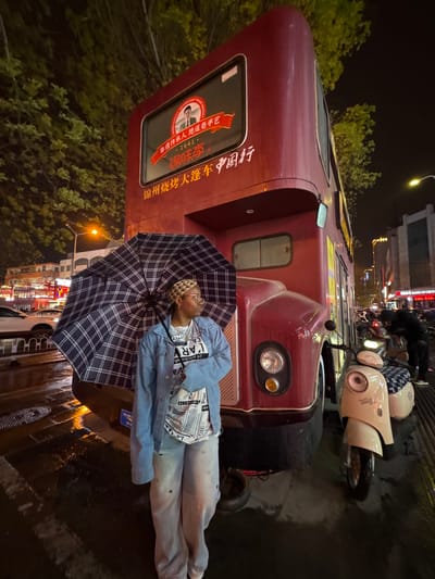 A person lies under a plaid umbrella next to a red bus on a wet street at night in Linghe, China. The bus has a sign that reads "锦州烧烤大篷车中国行" (JinZhou barbecue big tent car China tour) and "BANK OF CHINA". There are scooters parked nearby. Streetlights and buildings with lit signs are visible in the background. The person is wearing light-colored jeans, a patterned shirt, and a blue jacket. They also have a patterned head covering.