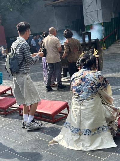 In a courtyard in Dongcheng District, China, on a paved surface with square tiles, a group of people are gathered. In the foreground, a person in a traditional ornate robe with intricate blue and white floral and bird patterns is kneeling on a red mat. Their elaborate headdress features gold ornaments and dangling embellishments. To the left, a young person wearing a striped long-sleeved shirt, light-colored culottes, white socks, and athletic shoes stands holding a wooden stick. They have a backpack on their shoulders. Behind them, other individuals are visible, including a person in a light-colored t-shirt and dark pants, and another in a patterned brown top and dark pants. A large, dark incense burner is present, emitting a plume of white smoke. In the background, a construction fence and scaffolding suggest ongoing work. Red ropes are strung across the scene, and stairs are visible to the right. The lighting indicates it is daytime, with sunlight casting shadows.