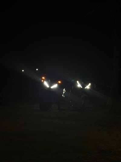Two motorcycles are parked on a dirt or gravel surface at night. The headlights of both motorcycles are illuminated, casting bright white light forward. The motorcycle on the left has its headlight in a V-shape. The motorcycle on the right also has its headlights on and appears to be a slightly different model, possibly with twin headlights. Both bikes have orange indicator lights visible. The background is dark and appears to be an outdoor environment, possibly a field or open area, with some faint lights visible in the distance. The lighting suggests a low-light or nighttime setting. The location is specified as Rawai, Thailand.