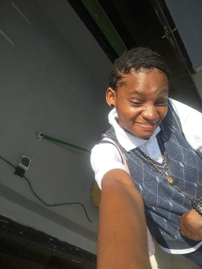 A young female, appearing to be a student, is captured in a head-and-shoulder selfie, positioned on the right side of the frame. She has dark skin, short braided hair, and wears small black stud earrings. Her eyes are squinted, and her mouth is open in a smile. She is dressed in a white short-sleeved polo shirt and a dark blue V-neck sweater vest with a light argyle pattern. A long gold-colored necklace featuring a medallion and a crucifix hangs around her neck, and a dark bracelet is visible on her left wrist. A light pink strap, possibly from a bag, is visible over her right shoulder. Her right arm extends into the foreground, holding the camera. The background consists of a plain light grey wall with a horizontal green pipe or conduit affixed to it. Below the pipe, a white electrical outlet with a dark square face is visible, from which a black cable extends downwards. Dark architectural elements are present along the top edge of the frame. The lighting is bright, illuminating her face. The specific location within Cotonou, Benin, cannot be determined from the provided visual information.