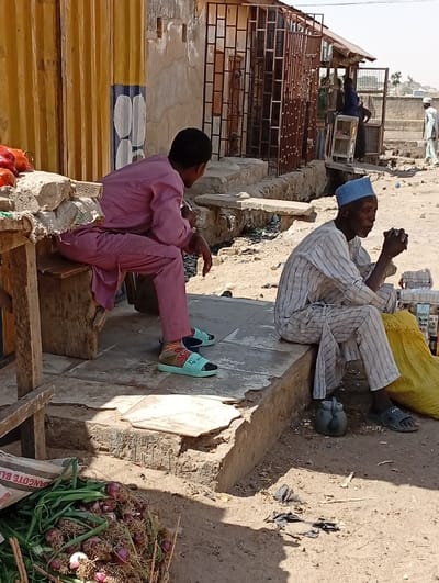 The image depicts an outdoor street scene in Gwagwarwa, Nigeria, under bright daylight. In the immediate foreground, a pile of green-stalked onions rests on the dry, light brown dirt ground, partially obscuring a white sack labeled "DANGOTE BL." To the left, a person wearing a pink, loose-fitting long-sleeved garment sits on a low wooden stool, facing away from the viewer and looking towards the right. This individual wears light blue slide sandals over patterned socks. Adjacent to them, on a rough wooden surface, is a cluster of red vegetables, possibly tomatoes or peppers. To the right, an older male with a dark complexion and a light blue cap is seated on a low concrete ledge. He wears a white-and-grey striped long-sleeved top and matching trousers, with dark open-toed sandals. His hands are clasped in front of him. A yellow bag rests near his left leg, and a small, dark, teapot-like object sits on the ground near his feet.
The ground throughout the scene is dry, light brown dirt, with scattered dark debris, including pieces of plastic. In the mid-ground, behind the seated individuals, structures include a corrugated yellow metal wall on the far left, a light-colored, weathered building wall featuring exposed metal grates over window openings, and a small, open-fronted stall-like structure with a corrugated metal roof. Concrete steps and ledges are visible near these structures. Several additional individuals are present in the mid-ground and background; one person is seen standing near a small display shelf, another in a light-colored outfit is partially visible, and a third person in white is walking towards the right in the far background. Distant buildings are vaguely visible under a hazy sky at the horizon.