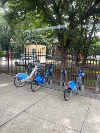 The image shows a row of blue and silver Citi Bike rental bicycles parked at a station on a sidewalk in New York City. The bikes are docked in a metal rack, and some are partially obscured by a black metal fence. Beyond the fence, lush green trees and foliage are visible, suggesting a park or tree-lined street. In the background, a light-colored building with dark vertical accents can be seen, along with a white car parked behind the fence. Sunlight filters through the trees, creating a dappled lighting effect. The scene appears to be set during the daytime, likely on a clear or partly cloudy day. The overall impression is one of an urban environment with readily available public transportation options. Text visible on the bikes includes "Citi Bike" and some numbers which appear to be a bike ID.