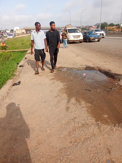 The image shows two young men walking side-by-side on a dirt road. The man on the left wears a checkered short-sleeved shirt and black shorts, while the man on the right wears a black long-sleeved shirt and dark pants. Both are wearing sandals. They are walking away from the camera towards the right side of the frame.
In the background, a busy intersection is visible with several vehicles, including a white van, a blue car, and a white SUV. There are also some people standing around and walking. The scene suggests a roadside area in Kagini, Nigeria.
The road is uneven and appears to be in disrepair, with a large puddle of water on the right side of the frame, containing a pink cup. There is also some litter on the road and on the grassy verge to the left. The lighting suggests it is daytime, likely late morning or early afternoon, with bright sunlight casting shadows. The sky is clear with a few scattered clouds. The overall impression is of a typical urban or semi-urban environment with signs of neglect.