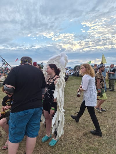 The image depicts an outdoor gathering, possibly a festival or event, in Lagunovo, Russia. The sky is filled with scattered clouds. In the foreground, a person wearing large white angel wings is standing, facing towards the right of the frame. To their left, a person with a black t-shirt and blue cargo shorts is partially visible. To the right of the person with wings, another individual in a white top and black pants is walking, carrying a dark bottle. Behind these central figures, a crowd of people and tents are visible, along with flags on poles. The ground is covered in dry grass.