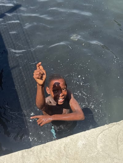 From an overhead perspective, a young Black boy is partially submerged in dark, murky water, looking up at the camera with a wide, joyful smile that reveals his teeth. His skin is wet and glistens under direct sunlight, which also creates bright reflections and speckles on the water's surface. His right arm is extended out of the water, with his hand curled and his index finger pointing towards the viewer. His left arm is also visible, with a glimpse of light blue-green fabric near his side, possibly part of his clothing.
To the right side of the image, a light-colored, textured concrete slab or edge of a structure is visible, suggesting the water body is man-made, possibly a canal or drainage ditch in Igboefon, Nigeria. The water contains small pieces of floating debris. The strong shadows and bright highlights indicate it is daytime with clear, sunny weather. The boy's expression conveys happiness and playfulness. No text is visible in the image.