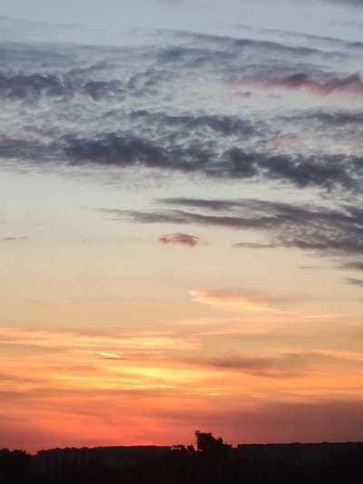 The image presents a stunning sunset over a silhouetted landscape, identified as Podolsk, Russia. The sky is the dominant subject, showcasing a vibrant and varied color palette.
In the upper section of the sky, soft pale blue hues transition into a light grey and white, adorned with textured, rippling clouds, characteristic of altocumulus or stratocumulus formations. These higher clouds exhibit subtle touches of pink and purple, reflecting the warm light from below. As the gaze moves downwards, the sky shifts to gentle beige and pale orange tones, featuring more scattered and elongated cloud wisps.
Closer to the horizon, the colors intensify dramatically. The sky bursts into brilliant shades of orange, deep red, and golden yellow, creating a fiery spectacle. These vibrant hues blend smoothly across the lower portion of the sky, with thin, elongated clouds brilliantly illuminated by the low-lying sun, appearing as streaks of bright light.
Along the very bottom of the frame, a dark, indistinct silhouette stretches across the horizon. This silhouette comprises buildings and what appear to be trees, indicating an urban or suburban environment. The dark foreground serves to highlight the dramatic light and color display in the sky.
The scene captures the tranquil beauty of dusk, with the sky serving as the main event. The weather appears partly cloudy, allowing for the interplay of light and shadow that creates the rich color gradient. There are no visible people, specific objects, or text within the image.