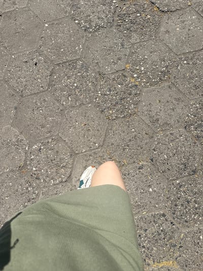 A first-person perspective shot captures a person's lower leg and foot as they stand on a textured, hexagonal-patterned pavement. The pavement is a dark gray concrete with visible aggregate and small debris, such as dried leaves and twigs, scattered across its surface. The person is wearing a light green, ribbed garment that covers their leg, and a white athletic shoe with teal accents. The shoe is positioned on the pavement, its sole visible. The lighting suggests it is daytime, with distinct shadows cast on the ground, particularly from the person's body. The scene appears to be outdoors, possibly a public walkway or plaza, given the paving style. The image provides no specific cues about the exact location within Haskovo, Bulgaria, beyond the pavement design. No text is visible in the image.