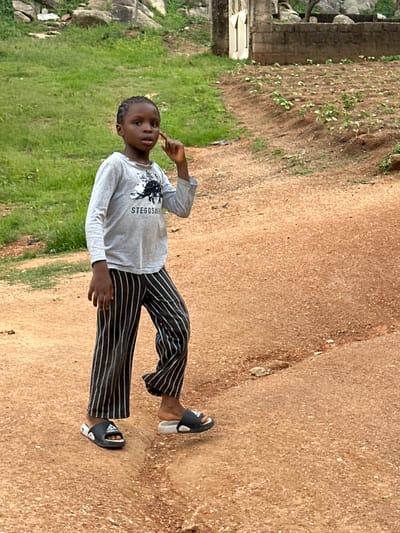 A young child is posing for a photo outdoors. The child is wearing a grey long-sleeved shirt with a black and white graphic and the text "STEGOSAURUS" printed on it, and black and white striped pants. The child is also wearing black slides with white stripes. The child is standing on a dirt path next to a grassy area. In the background, there are large rocks and what appears to be a small building. The lighting suggests it is daytime. The child's expression is neutral. The image was taken in Gwong, Nigeria.