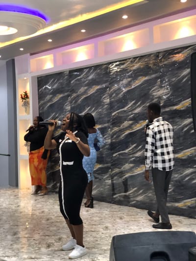 The image depicts an indoor performance scene in the city of Gbawe, Ghana. Four individuals are present on a polished, light-colored tiled floor with intricate patterns. The background features a large wall covered in dark, marbled paneling with white and gold veining. To the left, a lighter grey wall includes recessed shelving, one containing a white vase with red flowers. The ceiling is modern with recessed lighting, linear strip lights, and some purple and yellow accent illumination. A black speaker unit is partially visible in the bottom right corner.

In the foreground, a dark-skinned woman with long dark hair stands centrally, wearing a black knee-length dress with a white vertical trim and white accents, paired with white athletic shoes. She holds a black microphone to her open mouth, gesturing with her right hand, indicating singing or speaking. To her left and slightly behind, another dark-skinned woman wears a black long-sleeved top, an orange and yellow patterned skirt, and silver high heels. She also holds a black microphone to her mouth, appearing to sing. Further back and to the right of the central woman, a third dark-skinned woman wears a light blue, possibly lace, knee-length dress and black high heels, facing partially away from the camera. To the far right, a dark-skinned man with short dark hair stands holding a black microphone. He wears a black and white checkered long-sleeved shirt, dark grey trousers, and black dress shoes. All individuals appear to be actively participating in a group performance, with microphones suggesting a vocal presentation.