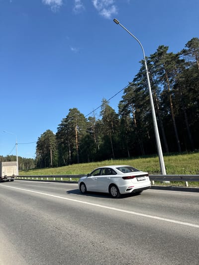 A white sedan, license plate O737YY 197, is traveling on a paved road with a white lane marker, alongside a forest of tall pine trees. A white truck is visible to the left of the sedan, also on the road. A silver metal guardrail separates the road from a grassy embankment. A tall street lamp with a curved arm is situated on the right side of the road, its pole casting a shadow. The sky above is clear and blue with a few wispy white clouds. The image appears to be taken in Koltsovo, Russia, likely on a sunny day.