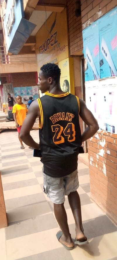 A young adult male, of dark complexion and short, dark, curly hair, stands angled slightly away from the viewer on a paved commercial walkway in Nnewi, Nigeria. He wears a black sleeveless basketball jersey with yellow trim, displaying "BRYANT" in yellow arched text above the number "24" in large yellow numerals on the back. He is also dressed in light grey cargo shorts and light grey slide sandals, with a dark mobile phone partially visible tucked into the back of his shorts.
The paved ground consists of alternating light beige and light grey stripes, running diagonally. To the main subject's left, a female in an orange t-shirt and reddish shorts stands further back, facing towards the right of the frame. Other indistinct figures are visible deeper in the background.
The immediate background includes a building facade with a prominent yellow sign featuring "CHUPÈZ COMMUNICATIONS" in blue lettering, identified as a mobile phone distributor in Nnewi, with additional partially visible text "TAMAGO COMMU... DISTRIBUTOR." Above this, a light blue section of the building has partial text, possibly "TECNO."
To the main subject's right, a reddish-brown brick wall, marked with patches of white residue, displays multiple light blue and white smartphone boxes. These boxes advertise "Infinix HOT 60 Pro+" and "Infinix SMART 10 Plus" models, some featuring the "MTN" logo. The scene is illuminated by bright daylight, casting visible shadows.