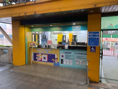 This media file captures an outdoor ticket booth, likely for a boat tour or ferry, given the visible body of water in the background. The booth features a counter with monitors and various signs, all housed under a yellow structural frame with a wooden ceiling. The ground in front appears wet, suggesting an overcast day or recent rain.

The main activity indicated is ticket purchasing and information dissemination. A large graphic on the counter advertises "游珠江, 小程序下单 立享优惠..." (Pearl River Tour, order via mini-program to enjoy discounts...). It also highlights "免出票" (Paperless/No ticket printing) options. Sections for "二维码购票" (QR code ticket purchase) and "电子票领取流程" (E-ticket collection process) are prominently displayed with QR codes and instructions.

A blue sign to the right of the counter lists departure times, with "08:00" and "18:40" clearly visible. Below this, a wheelchair accessibility symbol is affixed to a yellow column. Beyond the booth, metal railings lead to an area marked with a "禁止通行" (No Entry) sign, where a few individuals are visible in the background. The scene suggests a public transport or tourist facility in Guangzhou, China.