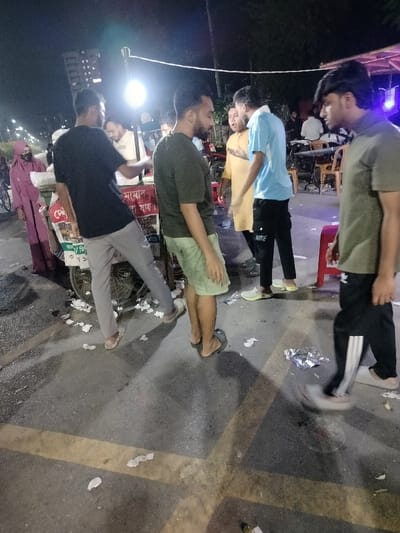 The scene takes place in a street market in Dhaka, Bangladesh, at the location marked by the coordinates 23.78 latitude, 90.374 longitude. A group of six men are standing in front of a food cart, engaged in conversation. There are several pieces of trash scattered on the pavement. The man on the left, wearing a black shirt and gray pants, appears to be talking to the man on the right, wearing a green shirt and black pants. The man in the blue shirt seems to be mediating the conversation. In the background, a woman in a pink hijab is walking away from the scene. The food cart is decorated with colorful signs and displays various food items. There is a motorcycle parked in the background. The street is paved and lit by streetlights.