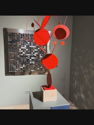 A red, abstract sculpture with circular elements is displayed in a gallery setting. The sculpture is suspended and composed of thin red rods supporting various sized red circles, some appearing flat and others slightly curved. It rests on a square, light-colored base. To the left of the sculpture is a large, framed artwork featuring a grid of small, reflective square and circular elements, creating a mosaic-like effect. The background is a plain grey wall. In the lower left corner, a white electrical outlet is visible on the wall. The overall scene suggests an art exhibition. The location is not New York City, United States of America.