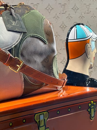 The image is a close-up, eye-level shot of a Louis Vuitton handbag with a patchwork design, featuring various textures and colors including the iconic monogram canvas, green fabric, and light grey fabric. A brown leather strap with a brass buckle is visible across the front of the bag. To the right of the bag is a mannequin head wearing a helmet with blue and white stripes and an orange and black racing suit, with a detailed profile of the face visible. Both the bag and the mannequin are placed on an orange, glossy surface, possibly a display case or the lid of a trunk, which has black and yellow decorative elements resembling hardware and clasps. The background is a light-colored wall embossed with the Louis Vuitton monogram pattern. There are no people present in the image, and no actions are being depicted. The scene appears to be set in a retail environment, likely a Louis Vuitton boutique, and based on the provided location, it is situated within Nong Prue Subdistrict, Thailand.