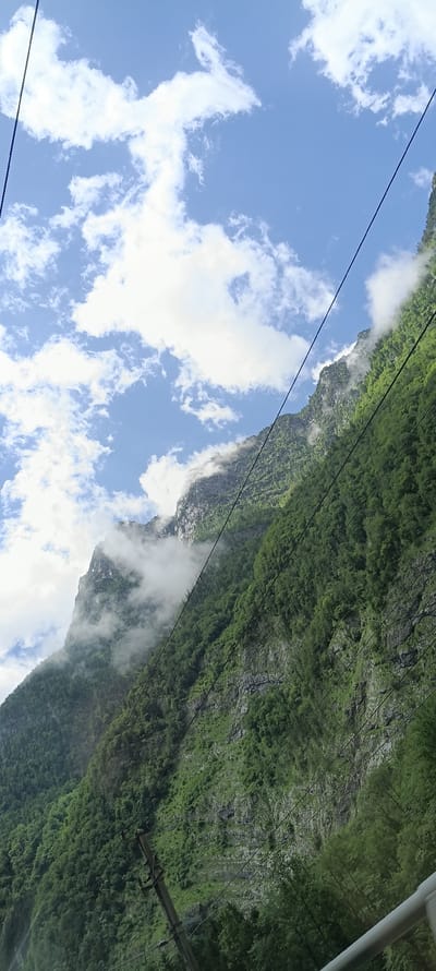 The image depicts a steep, verdant mountainside covered in dense trees, with rocky outcrops visible through the foliage. Wisps of white clouds cling to the upper reaches of the mountains, partially obscuring the rugged peaks. The sky above is a bright blue, dotted with fluffy white clouds. Several dark, thin wires crisscross the frame diagonally, suggesting the presence of a cable car system or power lines. In the lower portion of the image, a power pole with insulators is visible, further indicating utility infrastructure. The overall scene conveys a sense of natural grandeur and remote wilderness, with a touch of human industrial presence. The location is identified as Golling an der Salzach, Austria.