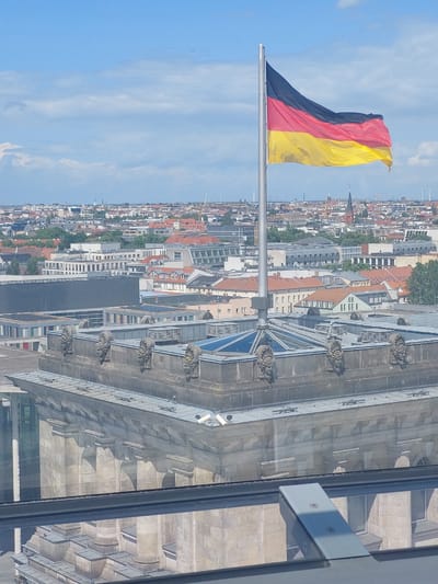 This is a high-angle, eye-level shot of the German flag flying from a flagpole, superimposed over a panoramic view of Berlin, Germany. The flag, a tricolor of black, red, and yellow, is prominently displayed in the center-right of the frame, billowing in the wind against a bright blue sky with scattered clouds.
In the foreground, the ornate top of a large, stone building is visible. The building's facade features classical architectural elements, including decorative sculptures of faces and columns. A large glass structure, possibly a dome or skylight, is visible on top of this building, reflecting the sky.
Beyond this building, the cityscape of Berlin unfolds. A dense collection of buildings with red-tiled roofs and various architectural styles stretches to the horizon. Distant church spires and modern structures, including wind turbines, can be seen in the background, indicating a mix of historical and contemporary development. The overall impression is one of a sprawling European capital on a clear, sunny day.