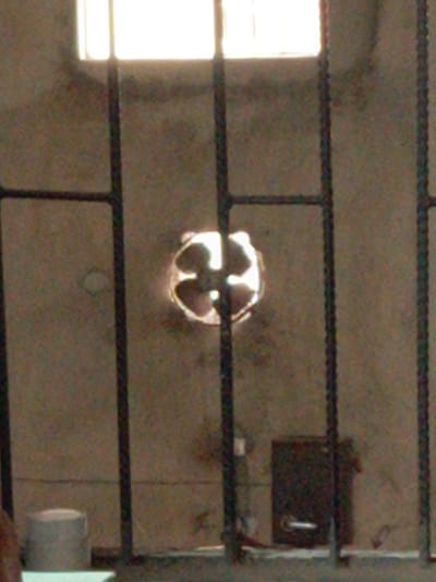 This image captures an interior view, likely of a secure or utilitarian room in Owerri, Nigeria, dominated by the presence of sturdy metal bars in the foreground.

At the top, a rectangular window emits a strong, bright light, indicating daytime. Below this window, the textured, unpainted concrete wall fills most of the background. Roughly in the center of the wall, there is a circular opening containing what appears to be an extractor fan or vent with visible blades, also illuminated from behind.

In the lower right section of the image, mounted on the wall, is a dark, rectangular box, possibly an electrical panel or a small utility box, featuring a distinct handle or latch on its front. To the lower left, a light-colored, cylindrical object rests on the floor or a low ledge. The bottom of the frame shows a horizontal bar or edge, likely part of the structure the metal bars are set into.

The scene conveys a stark and functional environment, with no people visible and no specific activity occurring. The rough concrete walls and the robust metal grille contribute to a sense of security or containment. No discernible text is visible in the image.