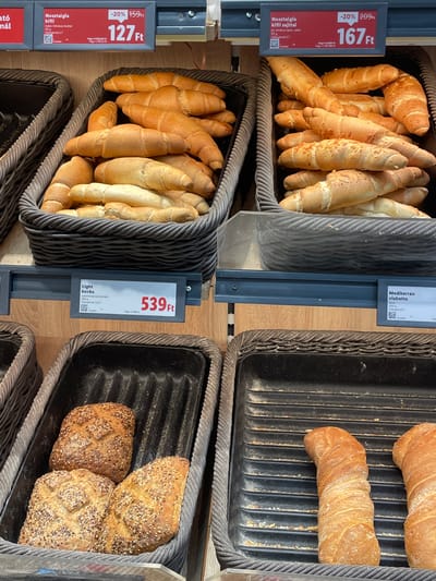 The image shows a display of baked goods in what appears to be a supermarket in Budapest, Hungary. In the foreground, there are several loaves of bread, some whole and some halved, in a dark basket. To the left, there are three loaves of bread with a textured, seedy crust. To the right of these, in a separate basket, are two long, twisted loaves of bread. Above these, in the middle of the image, are two more baskets filled with crescent-shaped rolls. The baskets are made of dark woven material. Above the baskets, there are price tags with Hungarian text and prices displayed in Hungarian Forint (Ft). One tag indicates a price of 127 Ft for "Nostalgia kifli," and another for "Nostalgia kifli sajtfal" shows a price of 167 Ft. Another price tag in the middle reads "539 Ft" for "Light kocka." A fourth price tag on the right shows "Mediterrán ciabatta." The overall environment is consistent with a bakery section of a retail store.