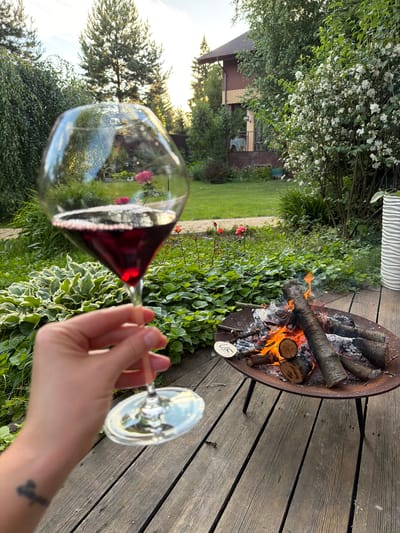 The image depicts a backyard scene in Zimyonki, Russia, on a wooden deck with a fire pit. A person's hand is visible, holding a wine glass filled with red wine. The wine glass is in the foreground, reflecting the surrounding environment, including a lush garden with green foliage and flowering plants, as well as a glimpse of a two-story house. In the middle ground, a fire pit contains burning logs and embers, with flames and smoke rising. The background shows trees and the sky, suggesting it is late afternoon or early evening. The overall atmosphere is serene and suggests a moment of relaxation in a domestic outdoor setting.