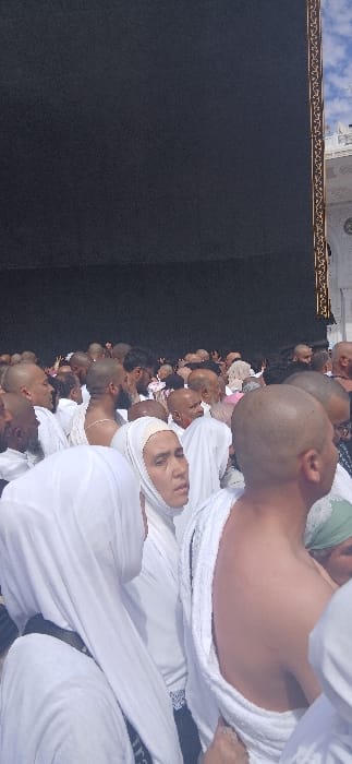 The image captures a dense crowd of pilgrims gathered within the Grand Mosque (Masjid al-Haram) in Makkah Al Mukarramah, Saudi Arabia, in proximity to the Kaaba. The upper portion of the frame is dominated by the dark black fabric (Kiswah) covering the Kaaba. A vertical section of gold-embroidered decoration is visible along the right edge of the Kaaba, adjacent to a white marble structure, part of the mosque's architecture, under a blue sky with scattered white clouds.
The foreground and midground are filled with numerous individuals. Many males are depicted wearing the Ihram attire, consisting of two seamless white cloths, with several having their right shoulder exposed. A significant number of these male pilgrims have shaved heads. Several females are also present, distinguished by their white head coverings (hijabs) and white garments. One woman in the central foreground is looking towards the right side of the frame. All individuals appear to be standing closely together in a high-density formation. The scene is illuminated by direct daylight.