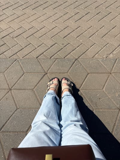 In Rusanovo, Russia, a person is sitting on a pavement. The pavement is composed of two sections with different patterns. The upper section has a herringbone pattern made of rectangular bricks, while the lower section features a pattern of interlocking hexagonal shapes. The person is wearing light blue jeans and beige sandals. Their legs are crossed, and their feet are positioned on the hexagonal pavement. A dark object, possibly a bag or a briefcase, is visible at the bottom of the image, partially obscuring the person's legs. The scene is illuminated by sunlight, casting a shadow to the right of the person.