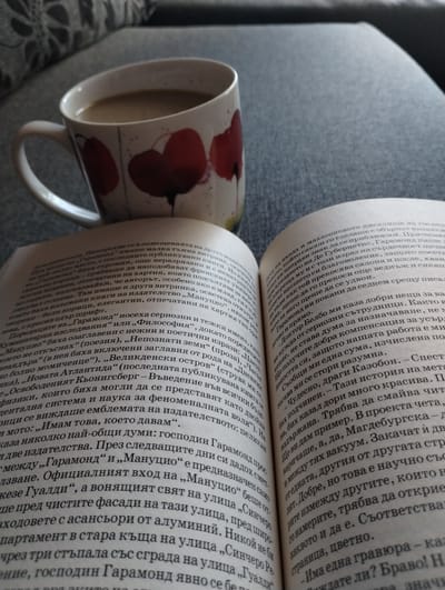 A close-up shot captures an open book resting on a gray, textured surface, with a mug partially obscuring the text on the left page. The book is filled with dense blocks of Bulgarian text, the pages slightly yellowed, with the binding curved open. The mug, predominantly white, is adorned with red poppy-like designs, containing a light-colored liquid, possibly coffee or tea. The gray surface appears to be a part of a couch. The text in the book appears to be about the publishing world. The photograph does not provide any information about the city of Yambol, Bulgaria.