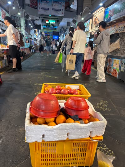 This image captures a street scene in the vicinity of the Seogwipo Maeil Olle Market in Seogwipo-si, South Korea. In the foreground, two large yellow plastic crates are stacked. The top crate contains a white styrofoam insert holding several red colanders filled with tangerines. A yellow basket sits on the ground in front of the crates, filled with red apples and peaches.
In the mid-ground, several individuals are walking through a covered market aisle. A man wearing glasses and a white t-shirt is looking to his left. A woman in a white t-shirt and jeans walks past, carrying a brown paper bag. A young girl in a pink jacket and red pants is walking ahead of her. A man in a grey hoodie and white pants walks in the background, looking to his right.
The background reveals a bustling market environment with various shops and illuminated signage. People are seen browsing and walking through the narrow passageways. The ground is paved with dark, textured tiles. The overall impression is of a lively, outdoor market scene.