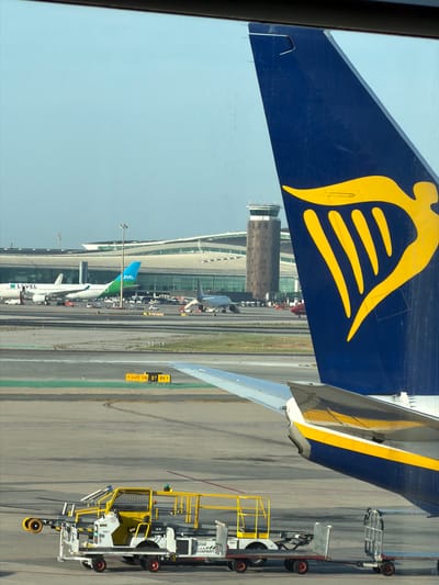 The image shows a view of an airport tarmac, likely from an airplane window. In the foreground, the tail of a blue airplane with a yellow Ryanair logo is prominent. The logo features a winged figure. To the left of the tail, the wing of the same airplane is visible, with a yellow stripe along its edge.
In the mid-ground, several other airplanes are parked on the tarmac. One is a white airplane with green and blue accents and the "LEVEL" logo. Another is a silver airplane. Various airport service vehicles are present, including a tug with a yellow cage structure and a flatbed trailer. In the background, an airport terminal building with a distinctive curved roof is visible, along with a control tower.
The scene is set at an airport, indicated by the airplanes, tarmac, terminal, and control tower. The presence of Ryanair's branding strongly suggests this is likely a Ryanair flight or at an airport where Ryanair operates. The location specified is El Prat de Llobregat, Spain, which is home to Barcelona-El Prat Airport (BCN), a major hub for airlines including Ryanair. Therefore, the scene is most likely at Barcelona-El Prat Airport. The weather appears clear with a bright sky. No people are clearly visible in the image.