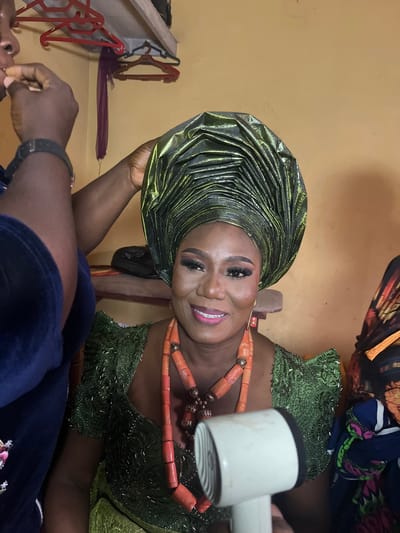 The image features a woman adorned in traditional Nigerian attire, suggesting a celebration or special event. She wears a green, intricately folded headwrap (gele) with a shimmering gold undertone and a matching green dress with beaded details. A coral beaded necklace is prominent around her neck. She is smiling and appears to be in the process of having her makeup applied or adjusted, as a hand holds a white object (possibly a makeup tool) near her face.
In the background, the wall is painted a pale yellow. Other individuals are partially visible, indicating she is likely surrounded by others preparing for the occasion. A clothing rack with hangers is visible on the wall. The image's specific location within Warri, Nigeria, cannot be determined solely from the visual information provided.