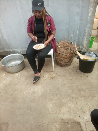 An adult female is seated on a white plastic chair, engaged in eating a meal from a white plate held in her left hand, while using a silver fork with her right. She wears a black Nike baseball cap, a dark reddish-brown open-front shirt over a black top, black leggings, and black open-toed slide sandals. Her hair is styled in long, light brown braids. To her immediate left on the ground is a large, circular, metallic pot. To her right, a large, cylindrical woven basket sits beside a black plastic bucket containing crumpled items. The background features a light grey wall with visible wear, and further right, several translucent yellow plastic containers and a green bucket are visible. The ground is a light-colored, patterned concrete or tiled surface. The scene is located in the city of Opete, Nigeria.