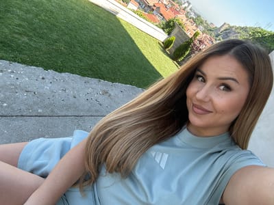 A woman with long, light brown hair is taking a selfie outdoors. She is wearing a light blue t-shirt with an Adidas logo on the left chest. Her hair falls over her shoulder and partially obscures her left arm. She is leaning on a rough, concrete surface, and behind her, there is a neatly manicured lawn. In the background, the cityscape of Asenovgrad, Bulgaria, is visible, characterized by red-tiled roofs and buildings situated on a hillside. The sky is clear and blue, indicating a sunny day. The overall scene suggests a relaxed, casual setting, possibly a garden or terrace overlooking the town.